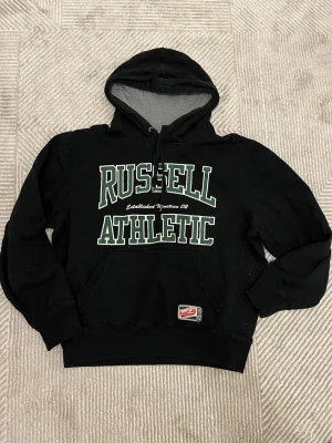 Svart hoodie Russell Athletic - Svart hoodie från Russell Athletic med stor vit och grön logga på bröstet. Klassisk huva med dragsko och magficka framtill. Insidan av huvan är grå. Perfekt för en sportig och avslappnad stil.