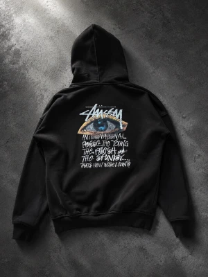 Svart Stüssy hoodie med grafiskt tryck - Säljer en svart hoodie från Stüssy med stort grafiskt tryck på ryggen och mindre tryck på bröstet. Trycket har ett öga och text i vitt och blått. Hoodien har huva och magficka, samt ribbade muddar vid ärmar och nederkant. Perfekt för streetwear-stilen.