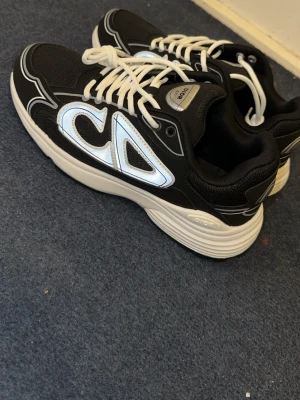 Svarta sneakers med vit sula och CD-detalj. Kommer utan box - Säljer ett par svarta sneakers med vit sula och stora reflekterande CD-detaljer på sidan. Skorna har vita snören och en chunky sula med svart och vitt mönster undertill. Materialet är en mix av mesh och syntet för en sportig look.