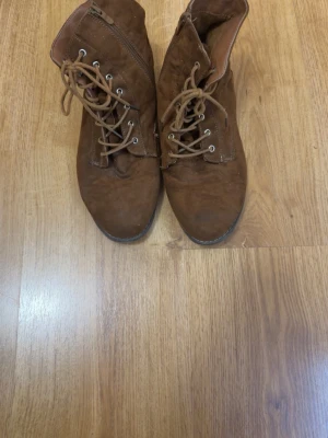 Bruna snörade boots i mockaimitation - Säljer ett par bruna boots med snörning och dragkedja på sidan. De har en klassisk rund tå, grov sula och är tillverkade i mockaimitation. Perfekta för dig som vill ha en stilren och enkel look till hösten eller vintern.