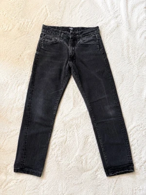 Svarta raka jeans med tvättad look - Säljer ett par svarta jeans med rak passform och tvättad finish. Jeansen har klassisk femficksdesign, bälteshällor och dragkedjegylf. Materialet är denim i bomull och de har en stilren, enkel look som passar till det mesta.