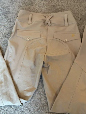 Beige ridbyxor från Maya delorez  - Snygga beige ridbyxor med helskoning i silikon för extra grepp. Byxorna har två bakfickor med knappar och bälteshällor i midjan. Smal passform och stretchigt material som ger bra rörelsefrihet. Perfekta för dig som gillar ridning och vill ha stilrena byxor.