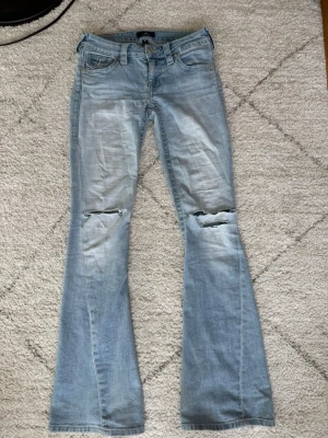 Ljusblå bootcut jeans True Religion - Säljer ett par ljusblå bootcut jeans från True Religion med slitningar och hål vid knäna. Jeansen har klassiska bakfickor med lock och dekorativa sömmar samt knappdetaljer. Perfekta för en avslappnad och trendig look.💕
