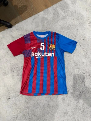 Barcelona matchtröja blå/röd - Säljer en Barcelona matchtröja i blått och rött med vertikala ränder, nummer 5 på både fram- och baksidan, samt klubbmärke och Rakuten-logga på bröstet. Tröjan har korta ärmar i olika färger och är tillverkad i ett lätt, ventilerande material. Ingen Dirket storlek 