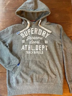Grå hoodie från Superdry - Snygg grå hoodie från Superdry med vit text på bröstet och stora dragsnören i huvan. Klassisk känguruficka framtill och mjukt material som känns skönt mot huden. Perfekt för en avslappnad streetstyle-look.