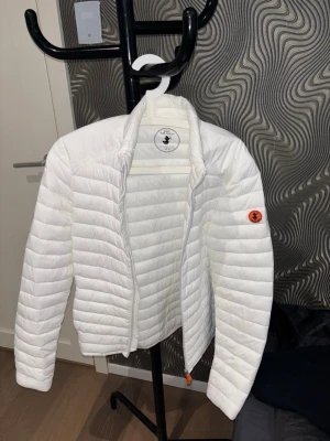 Vit dunjacka från Save The Duck - Säljer en snygg vit dunjacka från Save The Duck i storlek M. Jackan är quiltad med horisontella sömmar, har dragkedja framtill och en liten orange logga på ärmen. Tillverkad i lätt material som håller dig varm under vintern.