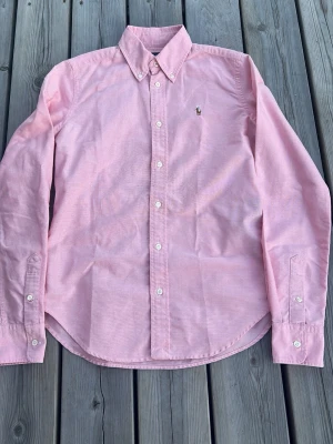 Rosa skjorta med knappkrage - Säljer en klassisk rosa skjorta med button-down krage och vita knappar. Skjortan har lång ärm och en broderad logga på bröstet. Tillverkad i mjuk bomull som känns skön mot huden. Perfekt för dig som gillar stilrena plagg med en twist.
