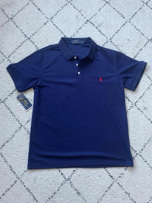 Marinblå pikétröja från Polo Ralph Lauren - Klassisk marinblå pikétröja från Polo Ralph Lauren med korta ärmar och krage. Tröjan har två knappar framtill och den ikoniska röda logotypen broderad på bröstet. Tveka inte att höra av dig vid frågor 😊