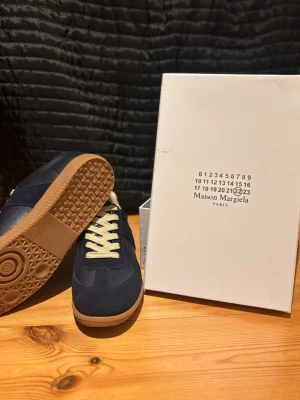 Maison Margiela gats - Snygga sneakers från Maison Margiela i mörkblå mocka och läder med beige snörning och klassisk gummisula. Ikonisk design med stilrena detaljer och låg profil som passar perfekt till streetwear eller casual outfits.