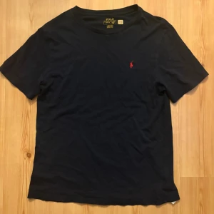 Mörkblå t-shirt från Polo Ralph Lauren - Snygg mörkblå t-shirt från Polo Ralph Lauren med klassisk rund hals och ikonisk röd broderad logga på bröstet. Tillverkad i mjuk bomull som känns skön mot huden. Perfekt för en stilren och avslappnad look.