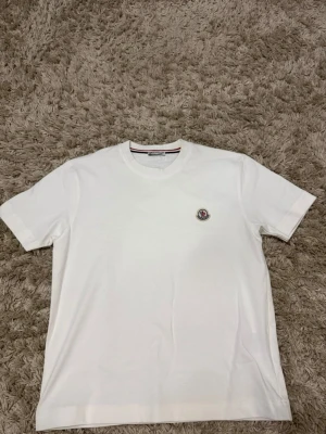 Vit t-shirt med liten logga - Klassisk vit t-shirt i bomull med rund hals och korta ärmar. På bröstet sitter en liten broderad logga som ger en clean och stilren look. Perfekt till jeans eller shorts för en enkel och snygg outfit.