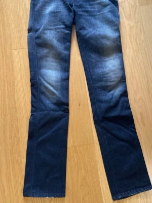 Mörkgrå bootcut jeans från Nudie - Snygga mörkgrå bootcut jeans från Nudie Jeans med klassisk femficksmodell och ljusa slitningar på låren. Jeansen har kontrastsömmar och vågig brodyr på bakfickan. Tillverkade i mjuk denim med skön stretch. Storlek 29/34