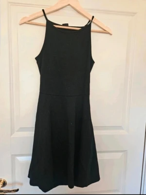 Klänning Gina Tricot S - En enkel och stilren svart klänning med smala axelband och rundad halsringning. Klänningen har en lätt utställd kjol och är ärmlös, vilket ger en clean och modern look. Perfekt för dig som gillar minimalistisk stil.  E4