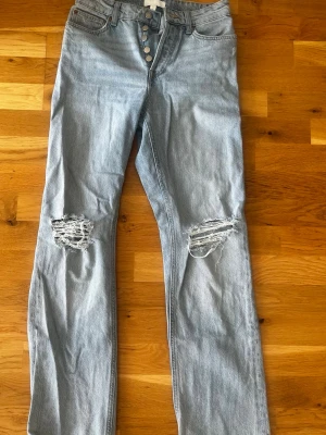 Ljusblå ripped jeans med knappar - Säljer ett par ljusblå jeans med raka ben och slitningar vid knäna. 