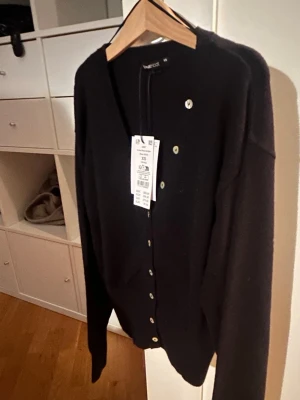 Svart kofta från Gina Tricot  - Stilren svart kofta från Gina Tricot i storlek XS. Koftan har v-ringning, ribbade muddar och knäppning framtill med silvriga knappar. Perfekt att slänga över en topp för en enkel och snygg look.