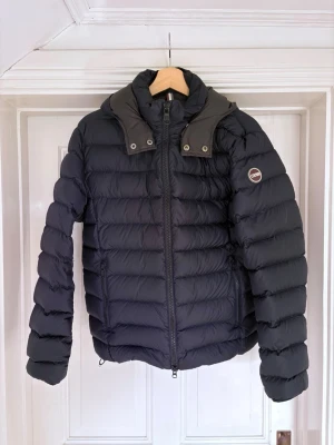 Colmar jacka S - Snygg svart dunjacka med avtagbar huva och dragkedja framtill. Jackan har två sidofickor med dragkedja och ett märke på ärmen. Puffig modell som håller dig varm under kalla dagar. Perfekt för dig som vill ha en stilren och funktionell jacka.