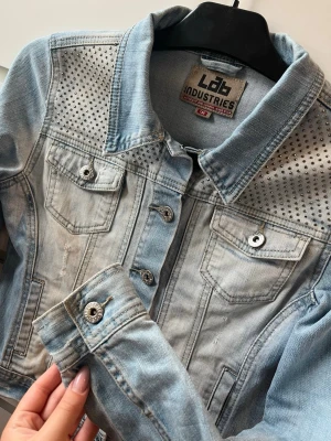 Ljusblå jeansjacka med nitar - Trendig ljusblå jeansjacka från Lab Industries med silverfärgade nitar på axelpartiet. Klassisk modell med två bröstfickor, knappar framtill och långa ärmar. Snygg tvättad look och detaljerade sömmar ger jackan en cool vibe. Kommer inte till användning. 