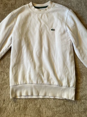 Vit sweatshirt från Lacoste - Klassisk vit sweatshirt från Lacoste med rund halsringning och den ikoniska gröna krokodilloggan broderad på bröstet. Tröjan har ribbade muddar vid ärmslut och nederkant. 7/10 skick, missfärgad krage.