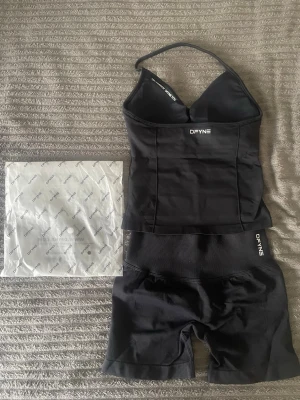 DFYNE impact set i svart - Säljer ett svart shaping-set från DFYNE med ett linne med smala axelband och vadderade kupor samt matchande höga shaping-shorts. Setet har en stilren design med logga på både linne och shorts. Materialet är stretchigt och följsamt för en bekväm passform.