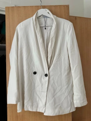 Vit oversized kavaj med svarta knappar - Stilren vit kavaj i oversized modell med två svarta knappar framtill. Kavajen har en mjuk, avslappnad krage och raka ärmar. 