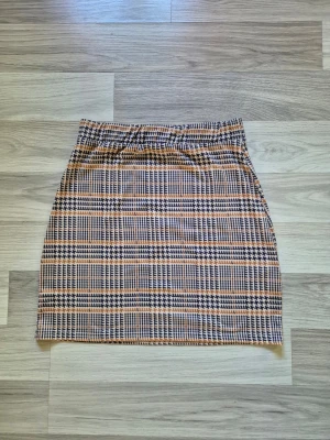 Rutig kortkjol i beige och svart - Säljer en snygg rutig kortkjol i beige, svart och brunt. Kjolen har en klassisk rak passform och elastisk midja för extra komfort. Perfekt att matcha med en enkel topp eller stickad tröja för en trendig look.