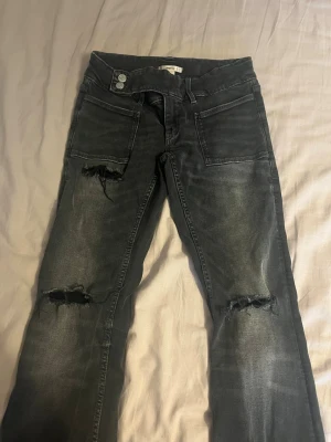 Mörkgrå bootcut jeans med slitningar - Säljer ett par mörkgrå low waist bootcut jeans med slitna detaljer över knäna och stora framfickor. Jeansen har utsvängda ben och en cool, tvättad look. Perfekta för dig som gillar en avslappnad och trendig stil.