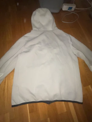 Nike Tech Fleece hoodie ljusgrå - Nike Tech Fleece hoodie i ljusgrå och vit med svarta detaljer. Dragkedja framtill, fickor på sidorna och en liten ficka på ärmen. Ikonisk Nike-logga i blått på bröstet. Tillverkad i mjukt och bekvämt material, perfekt för en sportig look.
