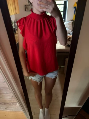 Röd blus med volangärmar - Snygg röd blus med hög smockad krage och volangdetaljer vid ärmarna. Blusen är ärmlös med lös passform och tillverkad i ett lätt material som passar perfekt till sommaren. Enkel att styla med både jeans och kjol.