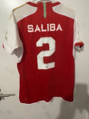 Arsenal matchtröja Saliba #2 - Röd och vit Arsenal matchtröja från Adidas med Saliba och nummer 2 på ryggen. Tröjan har klubbmärke, sponsorlogga och Champions League-märke på ärmen. Tillverkad i lätt och ventilerande material, perfekt för fotbollsträning eller match.