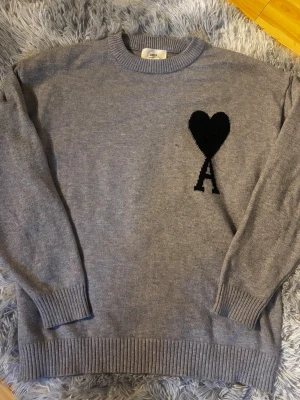 Grå stickad tröja från Ami - Snygg grå stickad tröja från Ami Alexandre Mattiussi med svart hjärta och bokstaven A broderat på bröstet. Tröjan har rund halsringning, ribbade muddar och är tillverkad i mjukt material. Perfekt för dig som gillar stilrena detaljer.