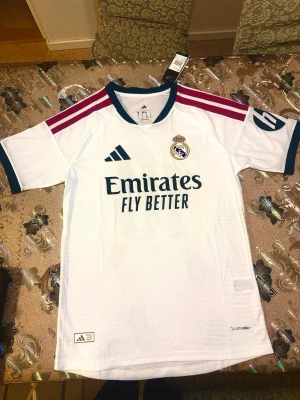 Real Madrid home Mbappé 10    26/27 - Säljer en vit Real Madrid matchtröja från Adidas med Mbappé och nummer 10 på ryggen. Tröjan har mörkblå detaljer, vinröda ränder på axlarna och klubbmärke samt sponsortryck på bröstet. Tillverkad i lätt och ventilerande material. 26/27 player version 