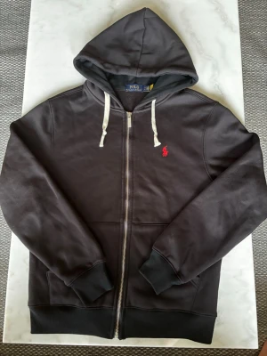 Svart hoodie från Polo Ralph Lauren - Snygg svart hoodie från Polo Ralph Lauren med dragkedja, vit dragsko i huvan och den klassiska röda loggan broderad på bröstet. Perfekt till dig nu när det börjar bli kallare.