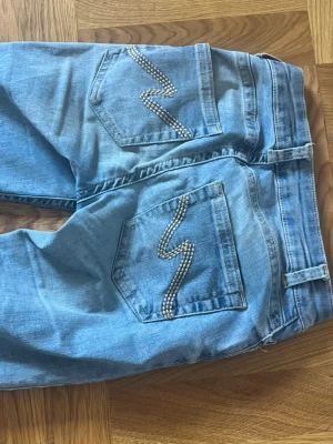 Ljusblå jeans med strass från Nelly - Snygga ljusblå jeans från Nelly med raka ben och dekorativa strassdetaljer i zigzag-mönster på bakfickorna. Klassisk femficksmodell i mjukt denimtyg, perfekta för en trendig och avslappnad look.