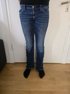 Blå bootcut jeans med broderade fickor - Snygga blå bootcut jeans med ljusa slitningar och kontrastsömmar. Baksidan har två fickor med coola broderade detaljer. Klassisk femficksmodell och knappgylf. Perfekta till vardagslooken och passar till det mesta.