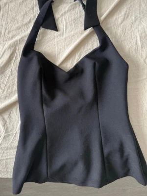 Svart halterneck korsettopp - Snygg svart korsettopp med halterneck och knytband i nacken. Toppen har en figurnära passform och markerad byst, vilket ger en stilren och trendig look. Perfekt att styla med jeans eller kjol för en cool outfit.