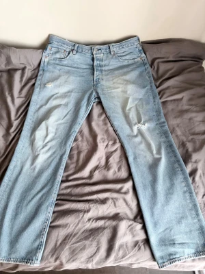 Levis 501 - Säljer ett par ljusblå jeans med raka ben och slitningar på låret och knät. Jeansen har klassisk femficksdesign och knappgylf. Materialet är denim i bomull och de har en avslappnad passform som passar till många olika stilar.