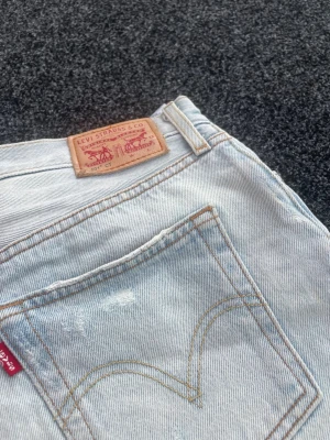 Ljusblå Levi's jeans - Ljusblå jeans från Levi's i klassisk femficksmodell med raka ben och slitna detaljer på båda benen. Jeansen har en vintagekänsla med synliga revor och Levi's ikoniska läderpatch bak i midjan. Perfekta för en avslappnad och trendig look.