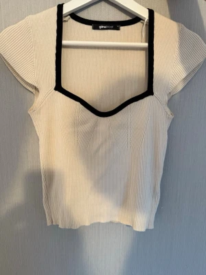 Ribbad topp med kontrastkant Gina Tricot - Snygg ribbad topp från Gina Tricot i ljus beige med svart kontrastkant runt den fyrkantiga ringningen. Kortärmad modell med figurnära passform och stretchigt material. Den har en skönt material och är perfekt till jeans eller kjol för den är enkelt att lyfta upp outfits.