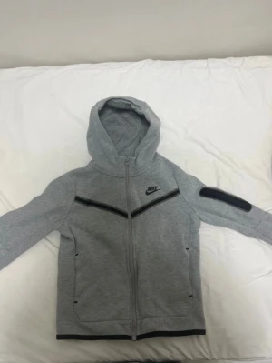 Nike tech kofta - Snygg grå hoodie från Nike med dragkedja framtill och svart logga på bröstet. Tröjan har huva, svarta detaljer längs bröstet och på ena ärmen samt fickor med dragkedja. Perfekt för en sportig och avslappnad stil.