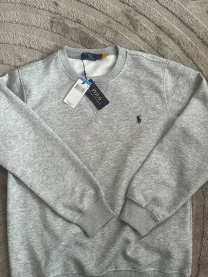 Grå sweatshirt från Polo Ralph Lauren - Grå sweatshirt från Polo Ralph Lauren med rund hals och marinblå broderad logga på bröstet. Tillverkad i mjukt bomullsmaterial och har ribbade muddar vid ärmar och nederkant för en klassisk och clean stil.