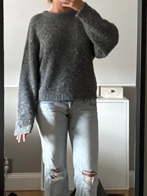 Mörkgrå stickad tröja med vida ärmar - Mysig mörkgrå stickad tröja med rund halsringning och extra vida, långa ärmar. Tröjan har en avslappnad passform och är gjord i ett mjukt, fluffigt material som passar perfekt för kyligare dagar.