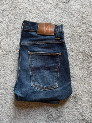 Mörkblå jeans från Nudie Jeans - Snygga mörkblå jeans från Nudie Jeans med klassisk femficksmodell och kontrastsömmar. Jeansen har en skinnpatch med logga bak i midjan och raka ben. Perfekta för en avslappnad och trendig look.