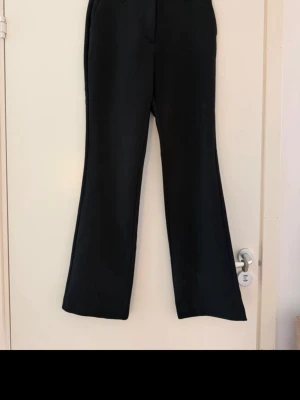 Svarta vida kostymbyxor - Snygga svarta kostymbyxor med hög midja och utsvängda ben. Klassisk design med fickor fram och passpoalfickor bak. Perfekta för en stilren look och passar till många olika outfits. Materialet ser ut att vara en mjuk och följsam blandning, troligtvis polyester eller viskos.