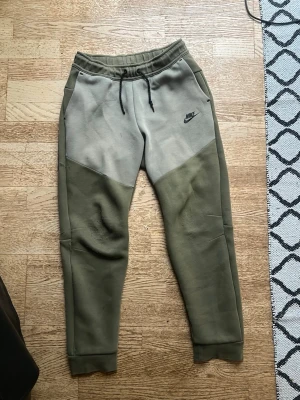 Nike mjukisbyxor i olivgrönt och beige - Nike mjukisbyxor i olivgrönt och beige med svart logga på vänster lår. Byxorna har dragsko i midjan, sidofickor och muddar vid bensluten. Tillverkade i mjukt material för maximal komfort och avslappnad stil.