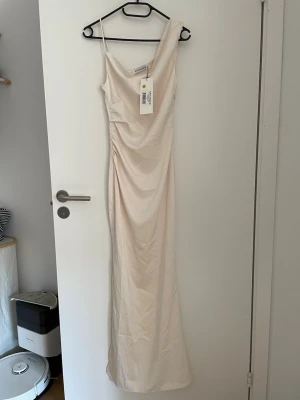 ADOORE almeria dress stl 32 - Elegant ljus långklänning med oneshoulder och smal passform. Klänningen har en draperad detalj framtill och är tillverkad i ett slätt, mjukt material. Perfekt för speciella tillfällen när du vill känna dig extra fin.