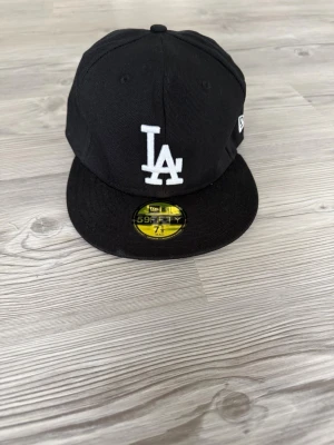 Svart LA New Era 59Fifty keps - Snygg svart keps från New Era med LA-logga broderad i vitt framtill och klassisk 59Fifty-sticker på skärmen. Kepsen har rak skärm och broderad New Era-logga på sidan. Perfekt för dig som gillar streetwear och sportig stil. 56.8 cm