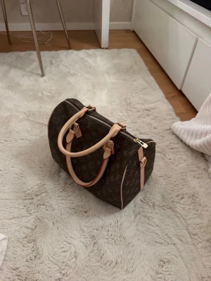 Väska - Säljer en klassisk handväska i brunt monogramcanvas med beige läderhandtag och detaljer. Väskan har guldfärgade metalldetaljer och dragkedja. Perfekt storlek för det viktigaste och ikonisk design. Kommer med axelband (EJ ÄKTA!!)