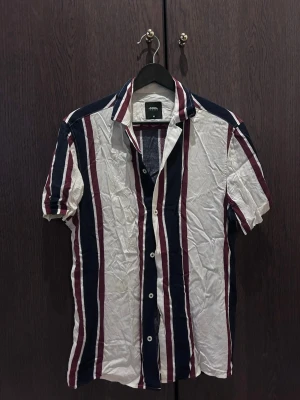 Randig kortärmad skjorta XS - Säljer en snygg kortärmad skjorta från BRITISH HERITAGE LONDON i storlek XS. Skjortan har vertikala ränder i marinblått, vinrött och vitt, klassisk krage och knäppning framtill. Perfekt för en avslappnad och trendig stil.
