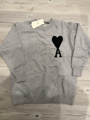 Ami sweatshirt - Snygg grå stickad tröja med svart hjärta och bokstaven A på bröstet. Tröjan har rund halsringning, långa ärmar och ribbade muddar vid ärmslut och nederkant. Perfekt för dig som gillar enkel men trendig stil.