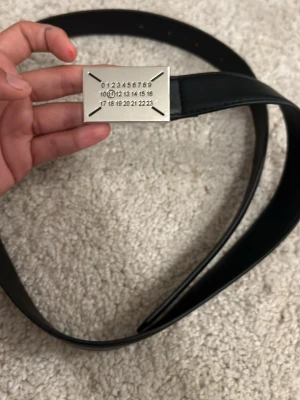 Maison marigela - Snyggt svart bälte med ett rektangulärt silverfärgat spänne som har siffror ingraverade i minimalistisk stil. Bältet har en clean look och passar perfekt till jeans eller kostymbyxor. Materialet känns lyxigt och spännet ger en unik touch.